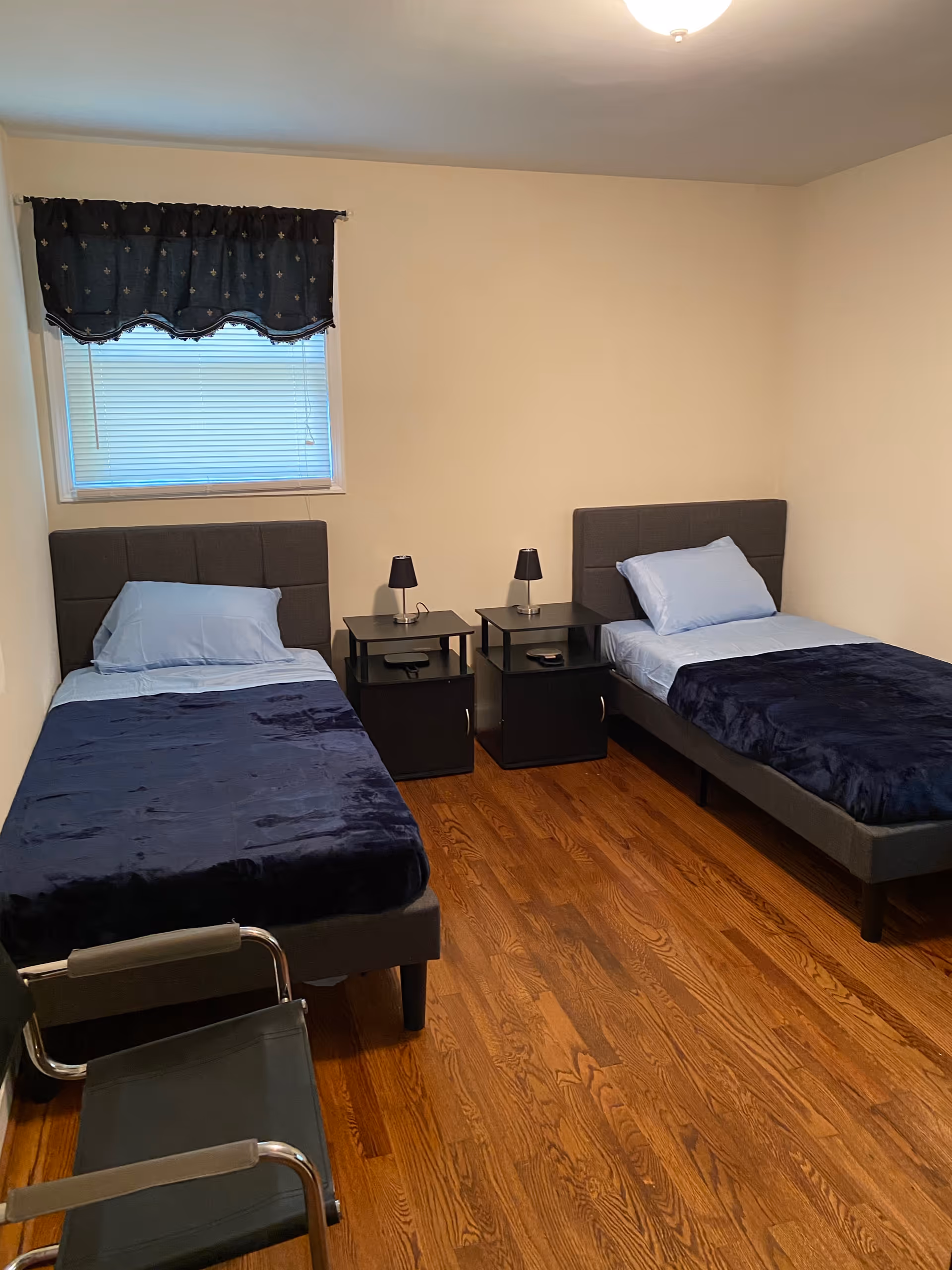 Simple bedroom with two twin beds, two nightstands with lamps, a window with a valance, and hardwood floors.
