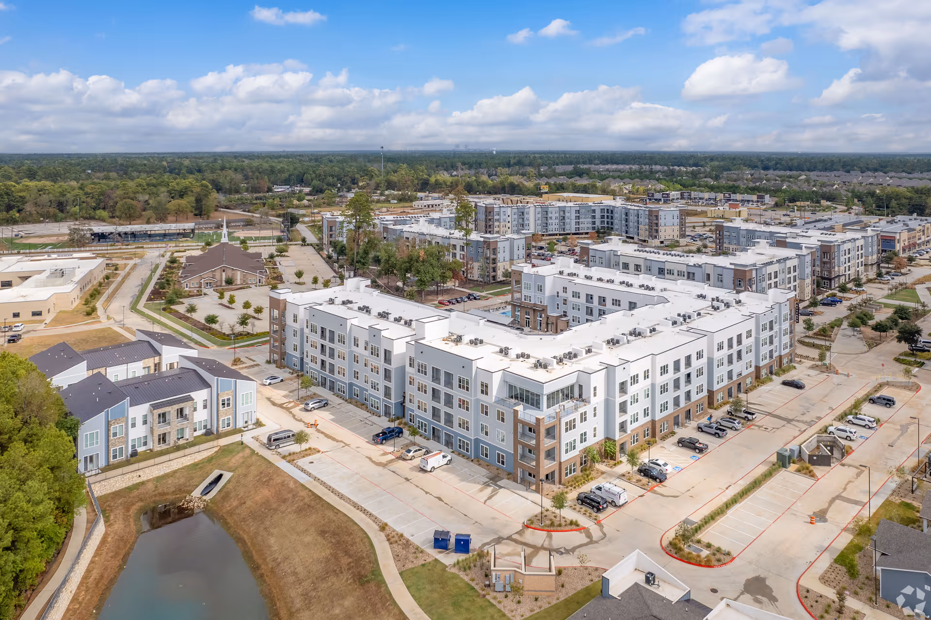 Aerial view of a large four-story senior living apartment complex with parking lots, surrounding buildings, and a small pond.