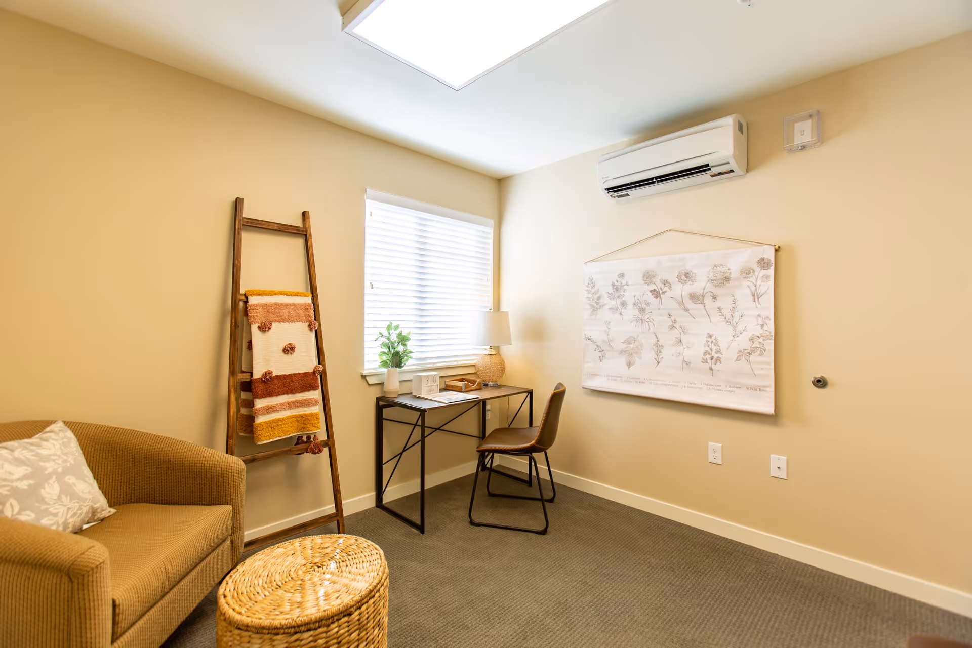 A cozy room with beige walls and carpeted floor featuring a small desk with a chair, a table lamp, a potted plant, and a decorative ladder holding folded blankets. There is a cushioned armchair with a patterned pillow and a woven round ottoman. A botanical print hangs on the wall beneath a mounted air conditioning unit.