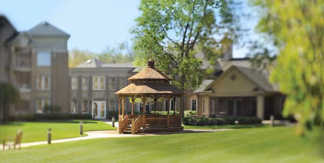 A wooden gazebo on a manicured lawn with residential buildings and trees in the background.