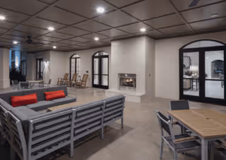 Indoor common area with modern seating including a sectional sofa with red cushions, a wooden table with chairs, rocking chairs near the wall, and a fireplace built into a white brick column. The ceiling has recessed lighting and the room features large arched glass doors.