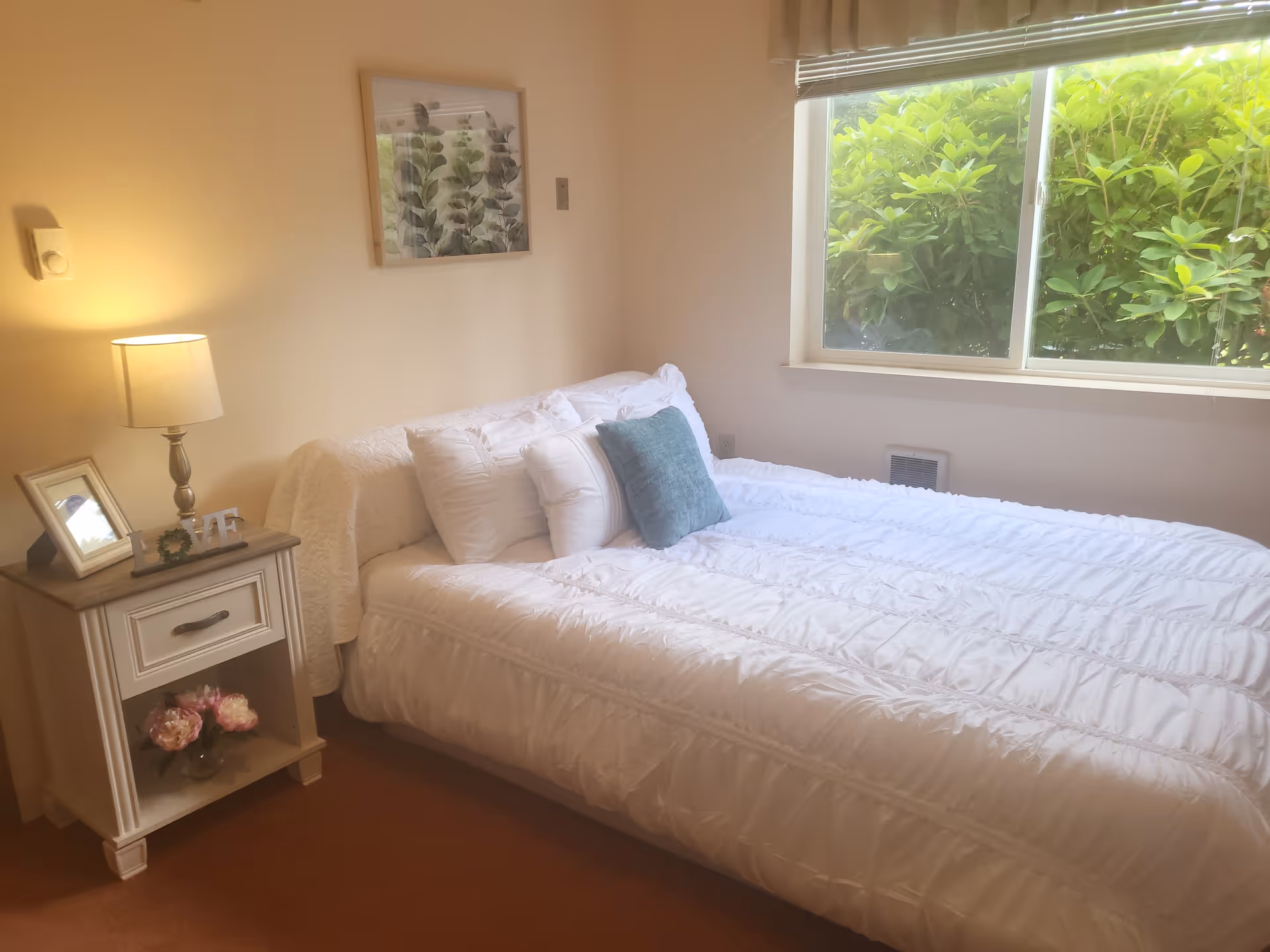 A cozy bedroom with a neatly made bed featuring white bedding and multiple pillows, including a blue accent pillow. Next to the bed is a small nightstand with a lamp, a framed photo, and decorative items. A window with a view of green bushes lets in natural light. A framed botanical print hangs on the wall above the bed.