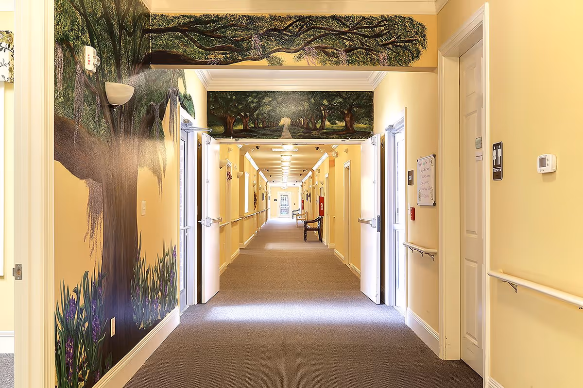 A long, well-lit hallway in a senior living facility with beige walls and carpeted floor. The walls feature a mural of large trees and greenery, creating a nature-inspired atmosphere. There are handrails along the walls, open double doors, and a bench further down the corridor. A restroom door is visible on the right side.