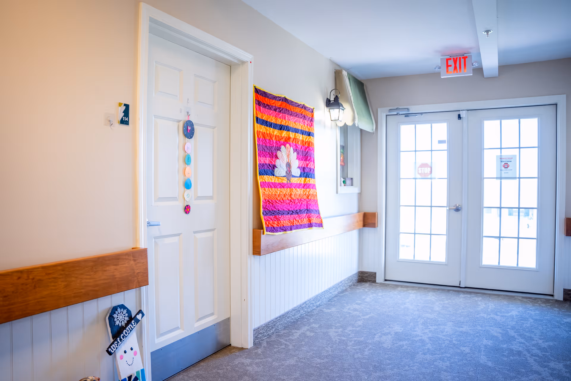 A bright hallway in a senior living facility with a white door labeled 114 on the left, decorated with colorful hanging ornaments. A vibrant quilt with red, orange, and purple stripes and a white flower design hangs on the wall. The hallway has light-colored walls, a wooden chair rail, and carpeted floor. At the end of the hallway, there are double glass doors with an exit sign above them letting in natural light.