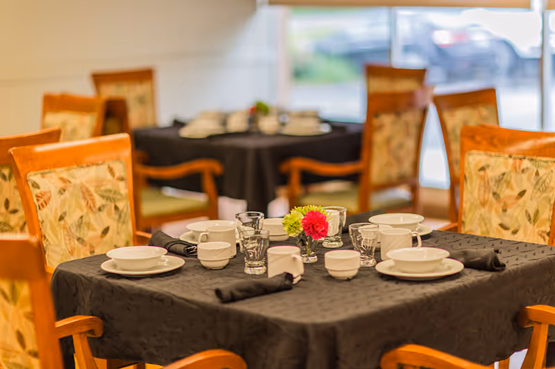 A dining room table set with plates, bowls, glasses, and a small vase of flowers surrounded by wooden chairs with floral upholstery.