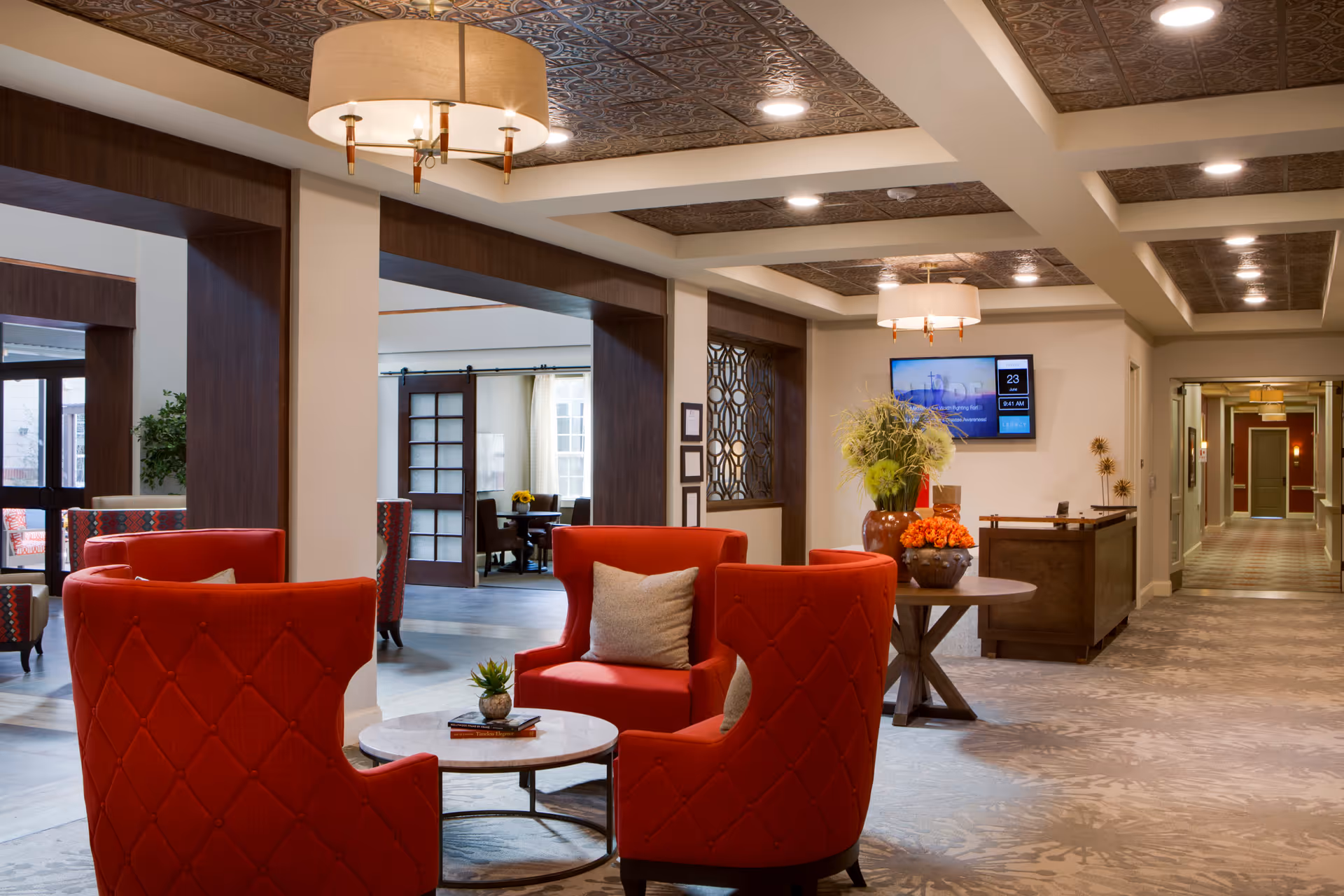 A spacious and well-lit senior living facility common area with red upholstered chairs arranged around a small round table. The ceiling features decorative panels and modern light fixtures. In the background, there is a hallway, a reception desk with floral arrangements, and a wall-mounted TV displaying information.