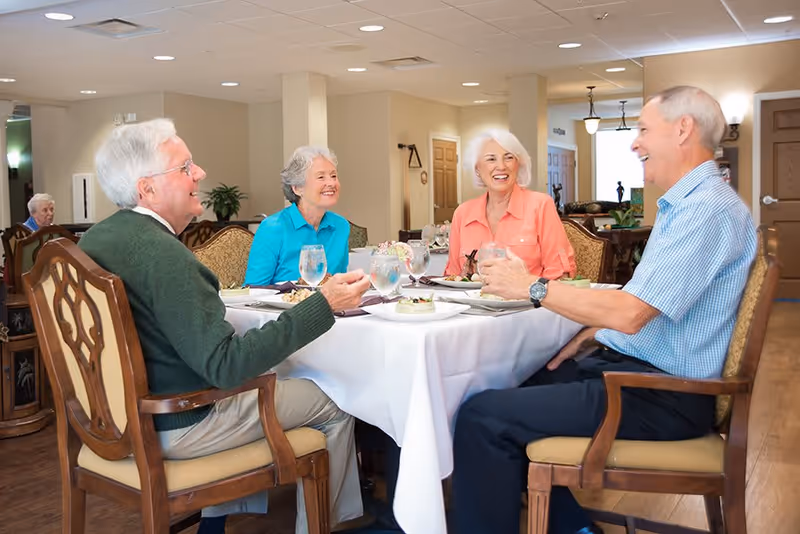 Four elderly adults sitting around a dining table in a well-lit room, smiling and enjoying a meal together with plates of food and glasses of water on the table.