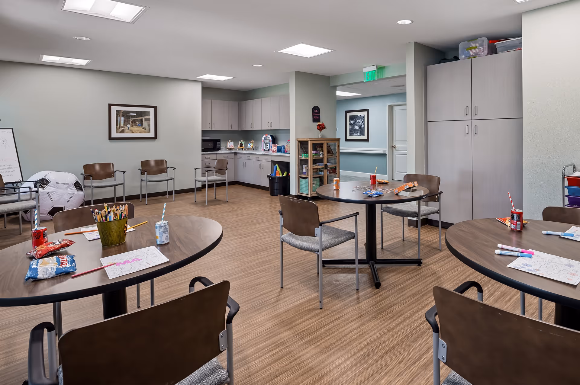 A well-lit activity room with round tables and chairs arranged around them. The tables have coloring sheets, markers, colored pencils, snacks, and drinks on them. In the background, there is a kitchenette with cabinets, a microwave, and a trash bin. There are additional chairs along the walls, a large soccer ball, a whiteboard, and a cabinet with games and supplies. The room has wood-patterned flooring and neutral-colored walls with framed pictures.