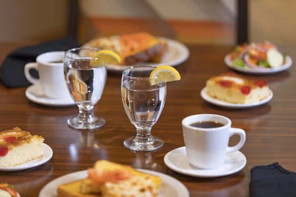 A dining table set with two glasses of water each garnished with a lemon slice, two cups of coffee on white saucers, plates with slices of cake topped with cherries, a small salad, and a sandwich in the background.