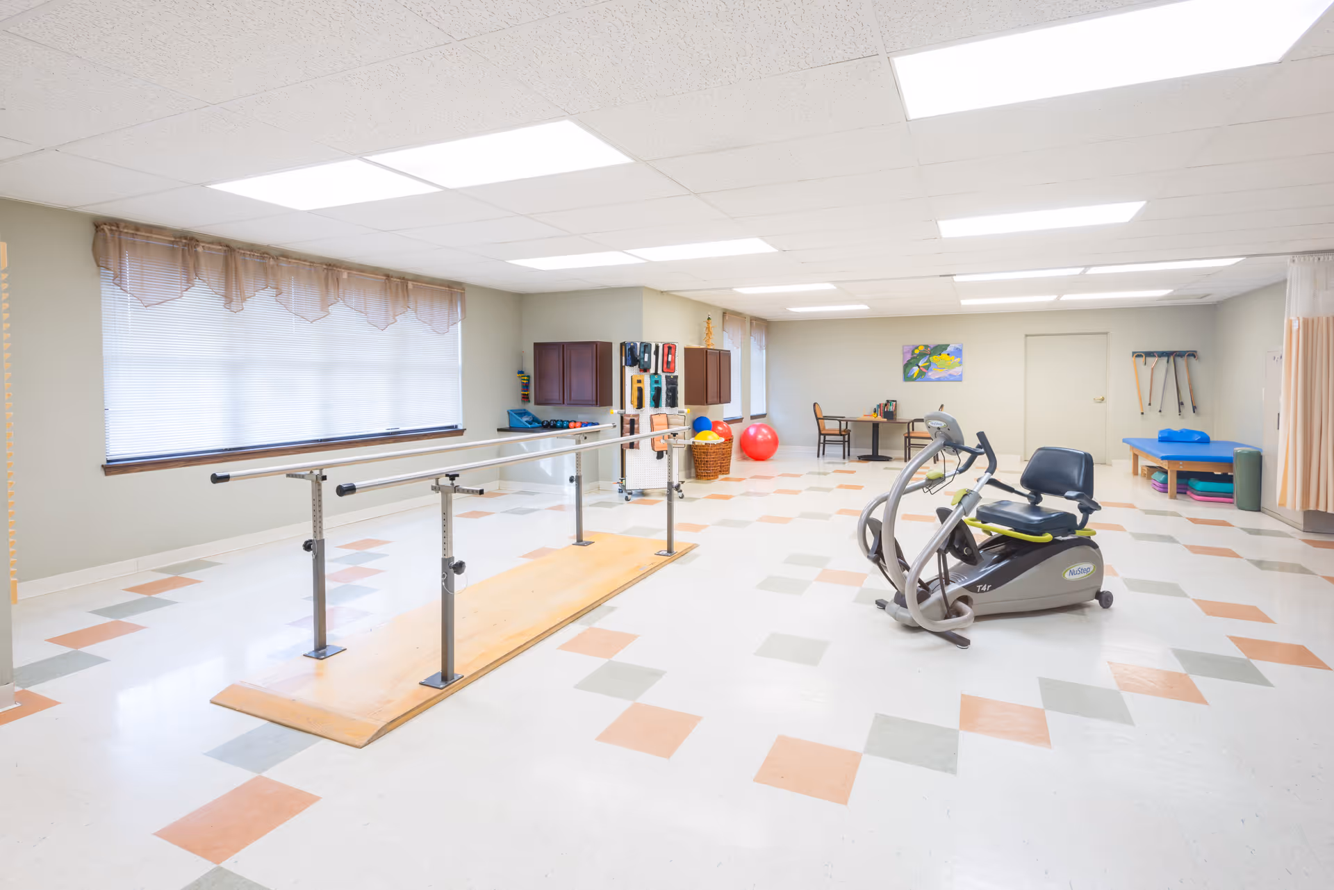 A spacious rehabilitation or physical therapy room with parallel bars for walking exercises, an exercise bike, therapy balls, and a treatment table. The room has large windows with blinds and curtains, tiled flooring with a pattern of orange and gray squares, and bright overhead lighting.