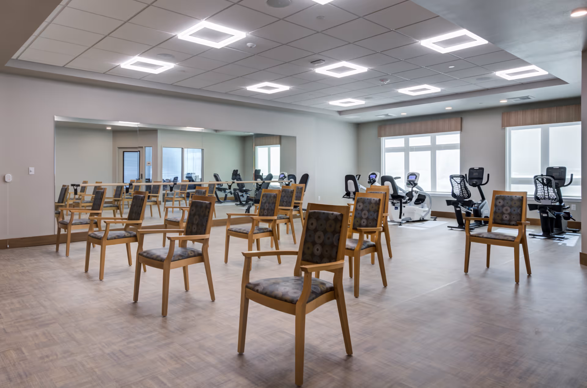 Spacious exercise/activity room with rows of wooden chairs, a mirrored wall and several recumbent exercise bikes by the windows.