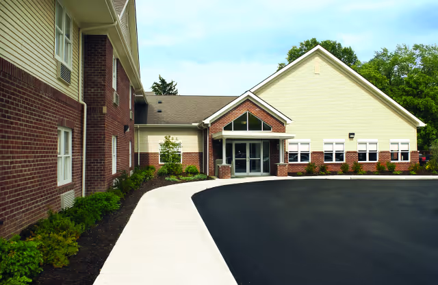 Exterior view of a two-story building with a combination of red brick and beige siding. The entrance features double glass doors under a small gabled roof with a triangular window above. There is a curved concrete walkway and a freshly paved black asphalt driveway in front of the building, surrounded by landscaped greenery and trees in the background.