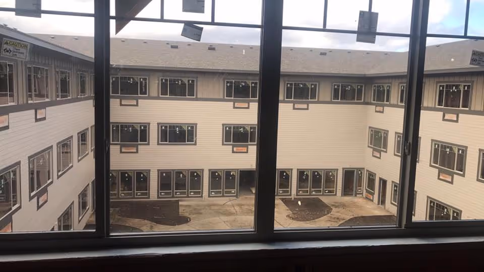 View through large windows showing the interior courtyard of a multi-story senior living facility building under construction, with beige siding and multiple windows on each floor.