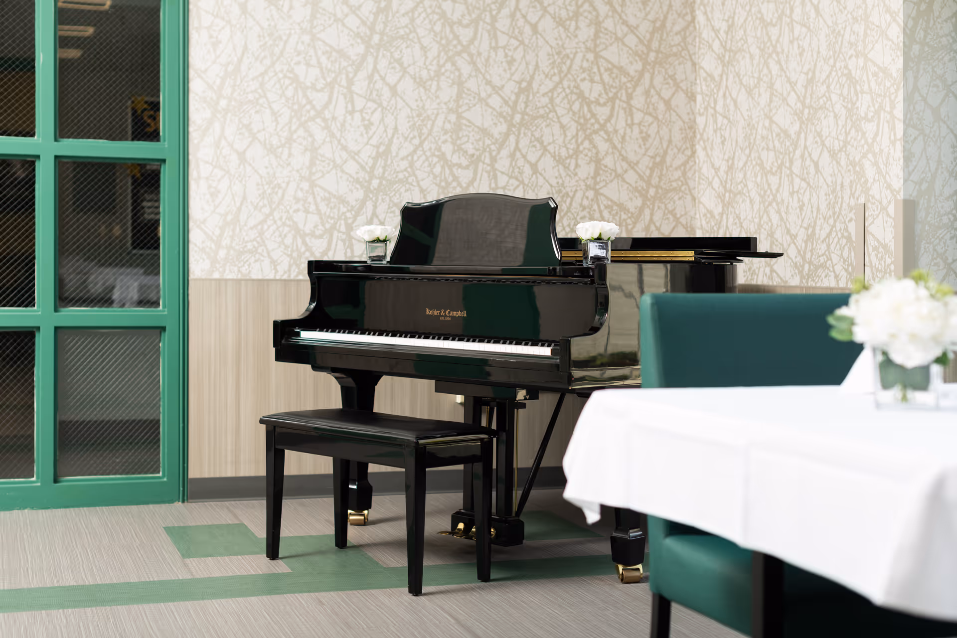 A black grand piano with a matching bench is placed against a wall with light patterned wallpaper. Two small flower arrangements sit on top of the piano. To the right, there is a dining table covered with a white tablecloth and green upholstered chairs. A green-framed glass door is visible on the left side of the image.