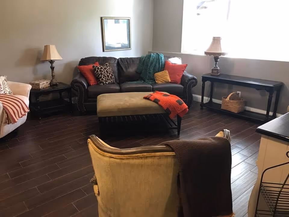 A cozy living room with a dark brown leather sofa adorned with colorful pillows and a green throw blanket. In front of the sofa is a beige ottoman with a red and black blanket draped over it. To the left, there is a beige armchair with a red and white striped throw. The room has dark wood flooring, a side table with a lamp, a framed picture on the wall, and a long console table with a lamp and a basket underneath near a large window letting in natural light.
