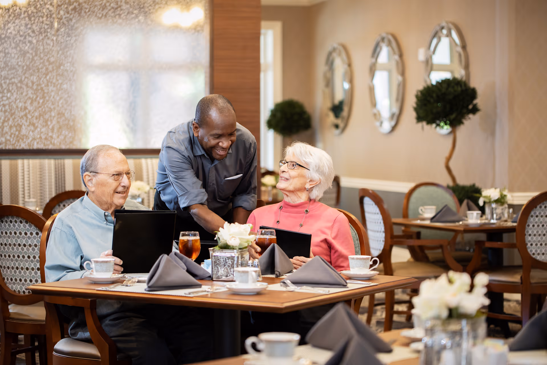A smiling waiter serves two elderly people seated at a dining table in a senior living facility. The table is set with cups, glasses of iced tea, napkins, and a small flower arrangement. The background shows a warmly decorated dining room with mirrors and potted plants.