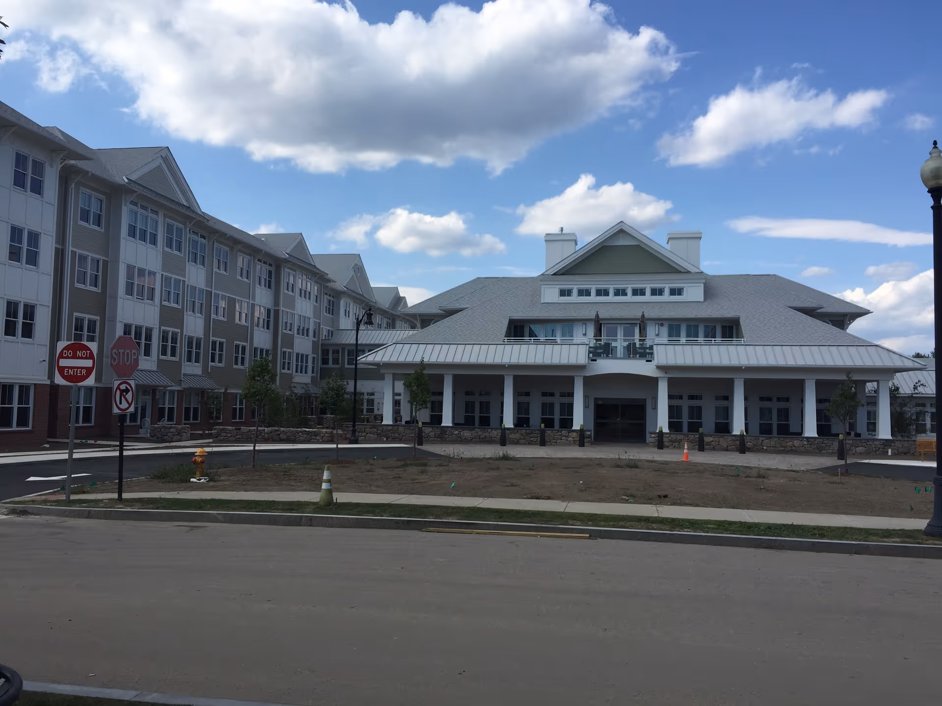 Exterior view of a large multi-story independent living community building under a partly cloudy sky. The building has multiple windows, a covered entrance with a balcony above it, and a paved driveway with traffic signs including stop and do not enter signs.