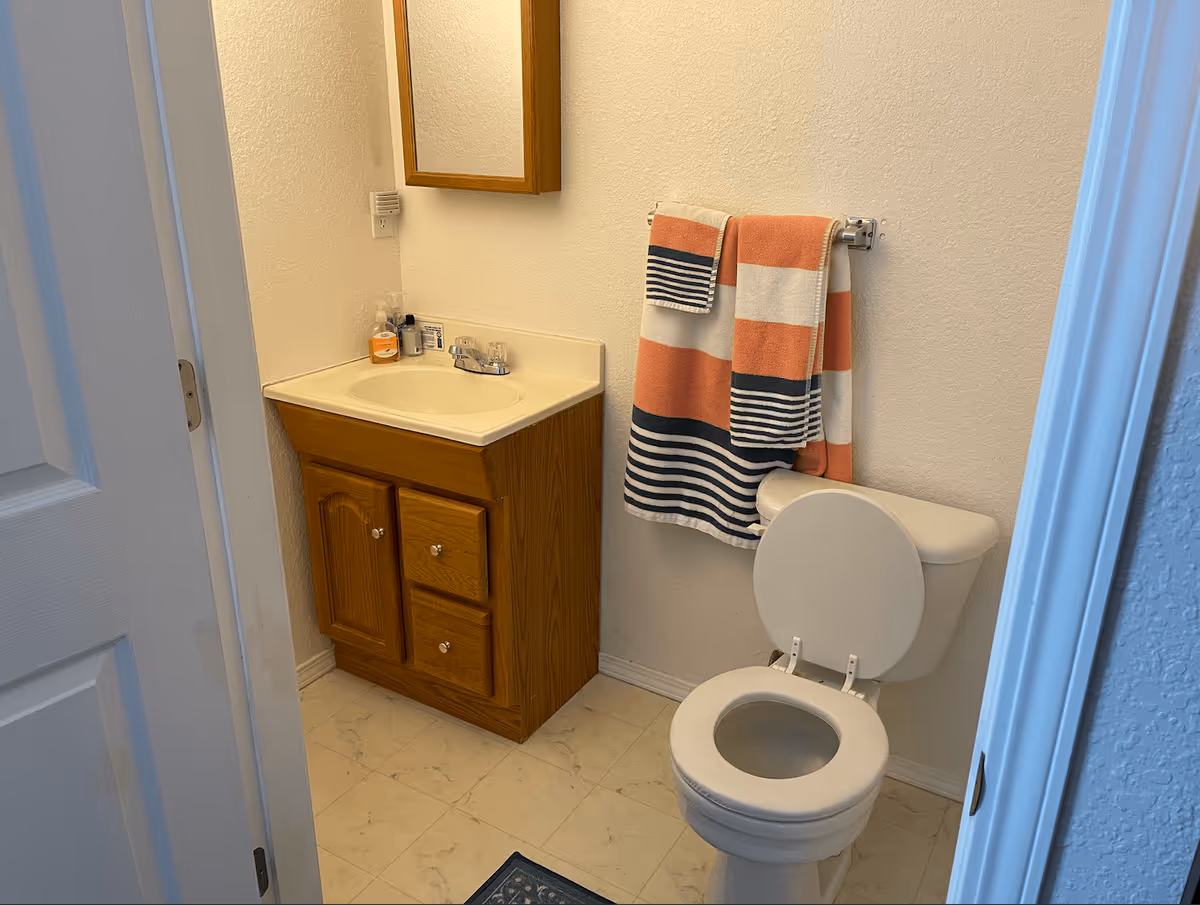 Small bathroom with a white toilet and a wooden vanity with a sink. Above the sink is a wooden framed mirror. Two striped towels in orange, white, and navy blue hang on a towel rack above the toilet. The floor has light-colored tiles and the walls are painted white.