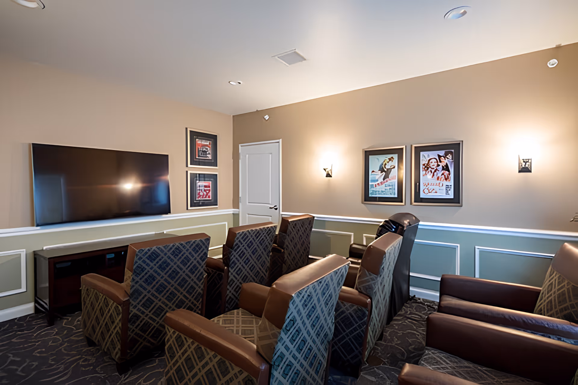 A cozy media room with several patterned armchairs arranged in rows facing a large flat-screen TV mounted on the wall. The walls are painted beige with white and green wainscoting, decorated with framed artwork and lit by wall sconces. A closed white door is visible in the corner.