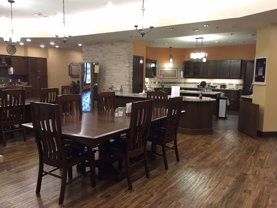 A spacious kitchen and dining area with wooden floors and dark wood furniture. The dining area features a large rectangular wooden table surrounded by eight matching chairs. The kitchen has dark wood cabinets, granite countertops, and modern pendant lighting. There is a refrigerator, a kitchen island, and various kitchen appliances visible. The walls are painted in warm beige and orange tones, and the ceiling has recessed lighting.