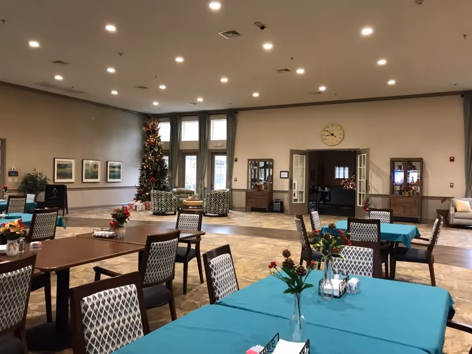 A spacious dining room decorated for the holidays with a Christmas tree in the corner. Several tables covered with teal tablecloths are arranged with chairs around them. Each table has a small floral centerpiece. The room has high ceilings with recessed lighting, large windows with curtains, and a clock above double doors leading to another room.
