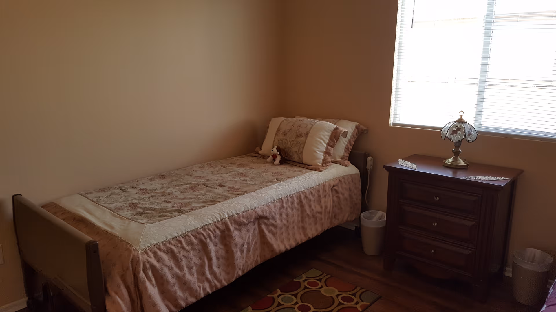 A small bedroom with a single bed covered in a floral patterned bedspread and matching pillows. Next to the bed is a wooden nightstand with three drawers, a decorative lamp, and a doily on top. The room has beige walls, a window with blinds letting in natural light, and a small colorful rug on the wooden floor.