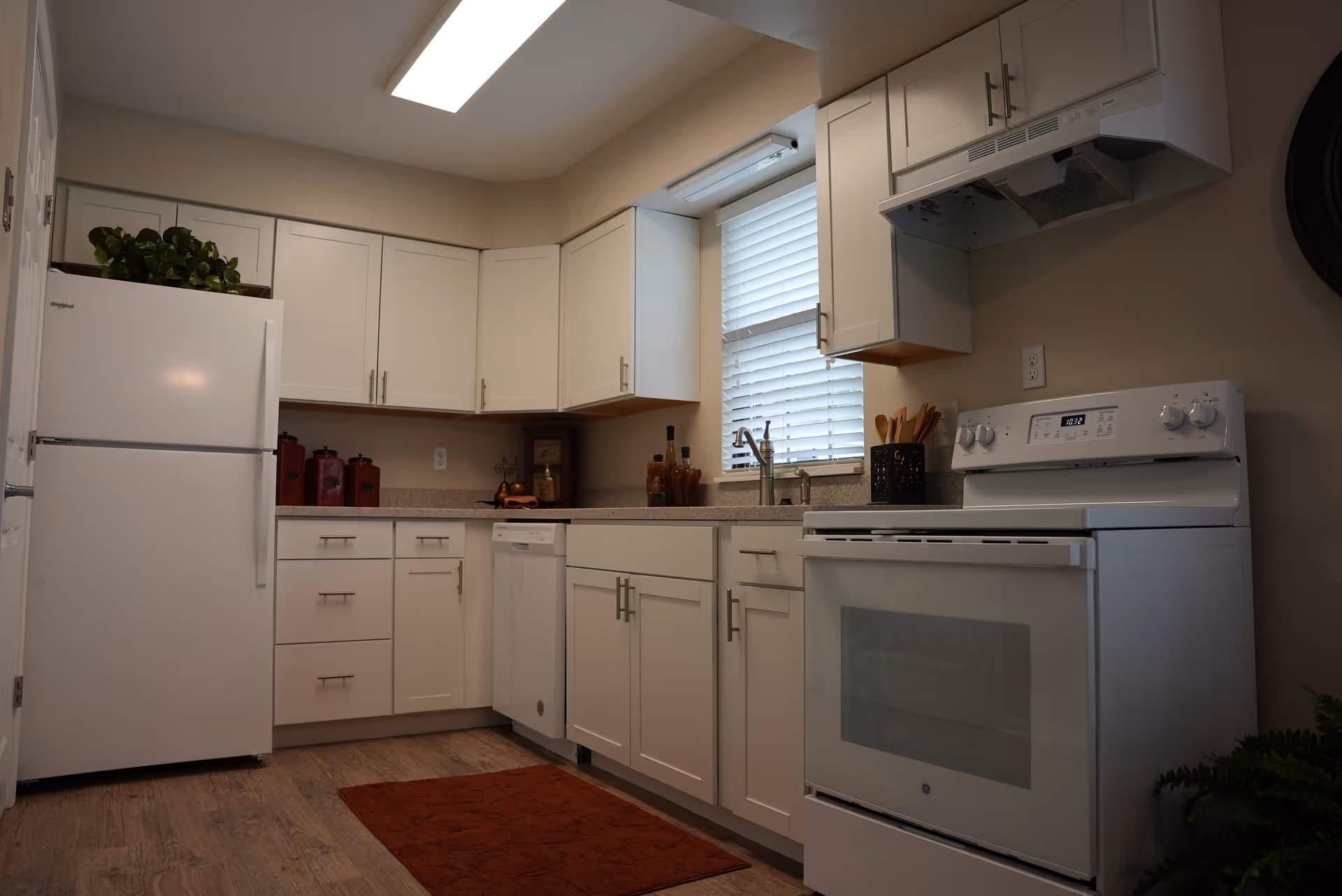 A clean and modern kitchen with white cabinets, a white refrigerator, a white stove with an oven, a dishwasher, and a window with blinds above the sink. There are decorative items on the countertop and a red rug on the wooden floor.