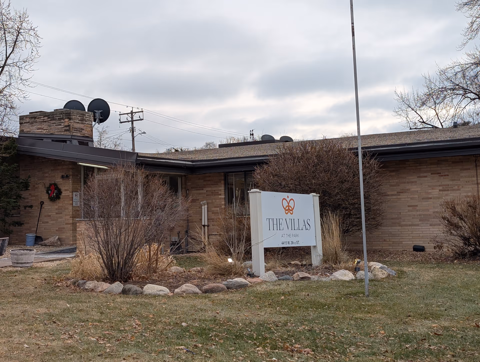 Exterior view of a single-story brick building with a sign in front that reads 'THE VILLAS AT THE PARK 4415 W. 36 1/2 ST.' The building has a chimney with satellite dishes on top, leafless bushes, and a grassy area with scattered leaves. The sky is cloudy.
