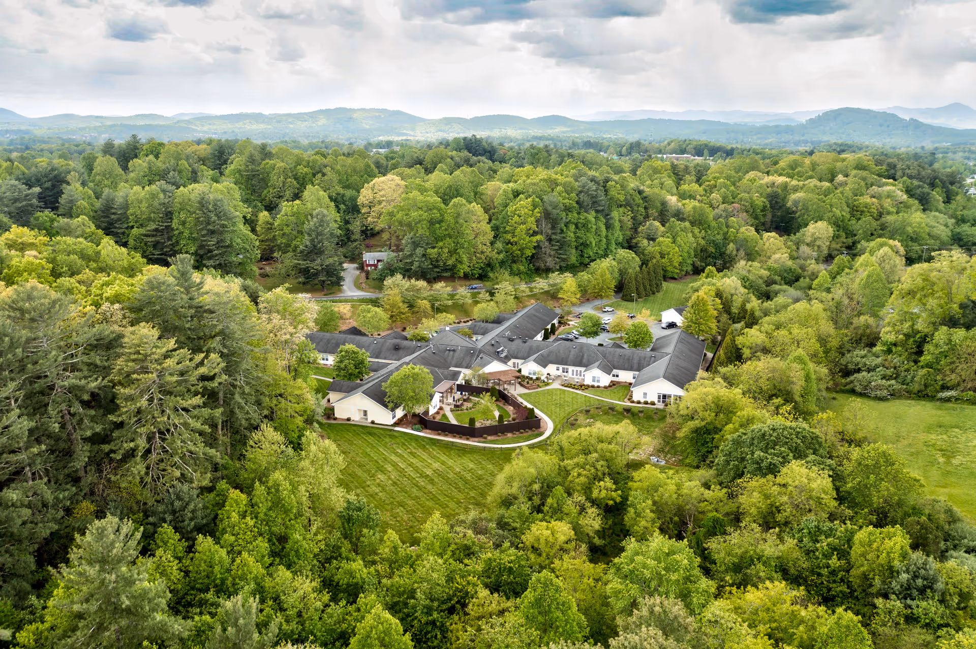 Aerial view of Carillon Assisted Living of Hendersonville surrounded by dense green trees and rolling hills under a cloudy sky.