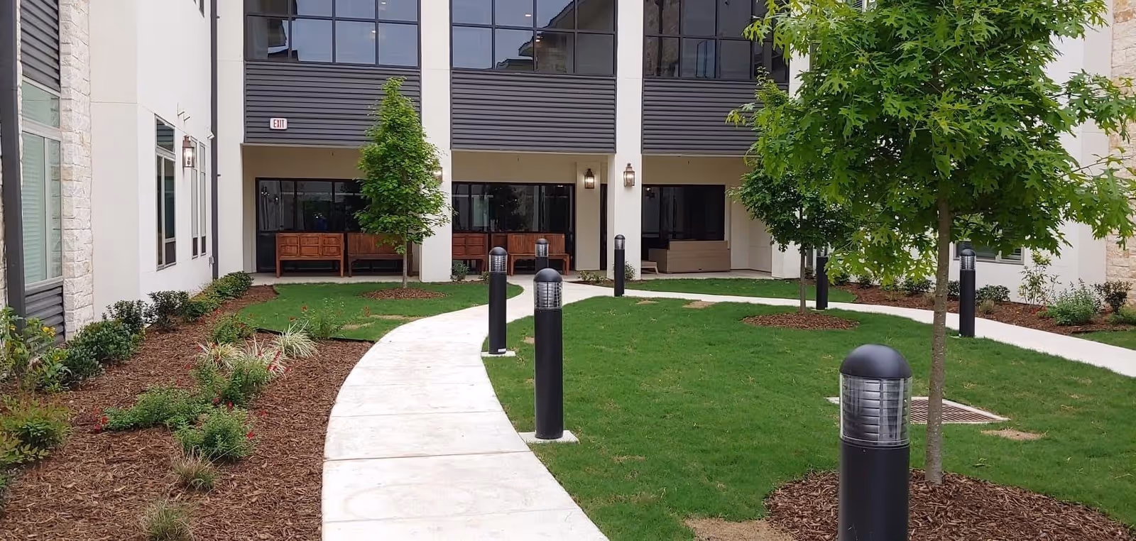 Outdoor courtyard area with a curved concrete walkway, green grass, young trees, and black cylindrical outdoor lights. The building exterior features large windows, beige walls, and stone accents. There are wooden benches and a seating area under a covered section of the building.
