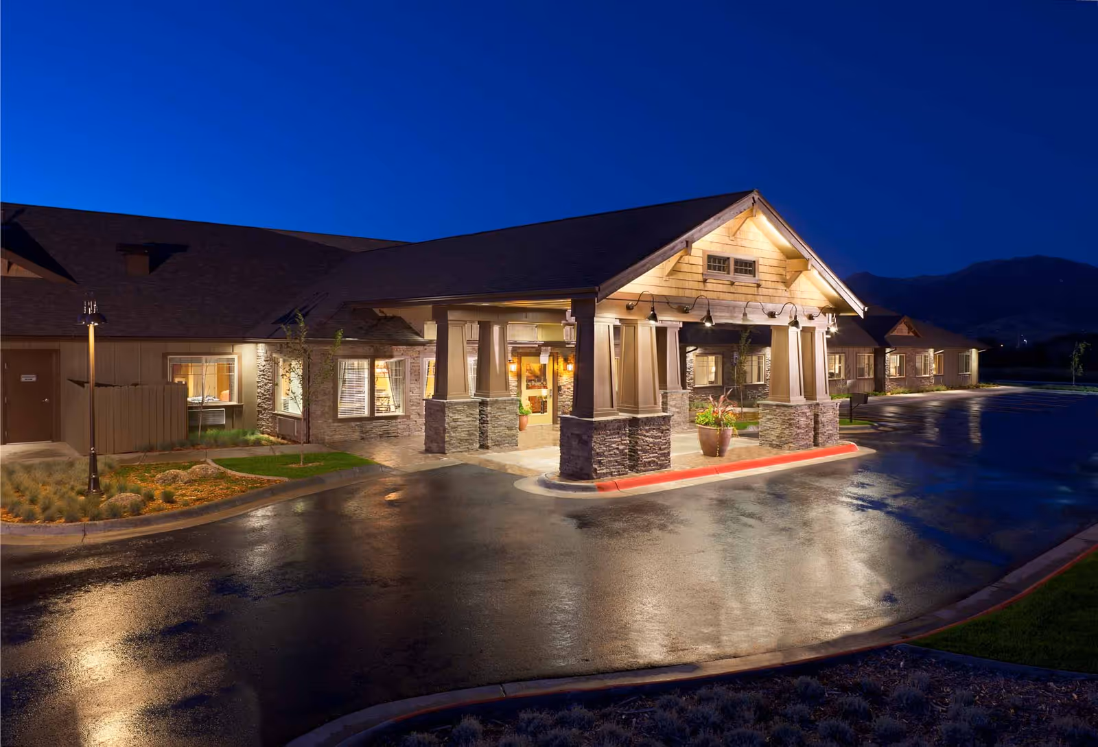 Exterior view of Spring Creek Inn Memory Care Community at night, showing the well-lit entrance with stone pillars and a covered driveway, surrounded by a wet driveway and landscaped greenery.