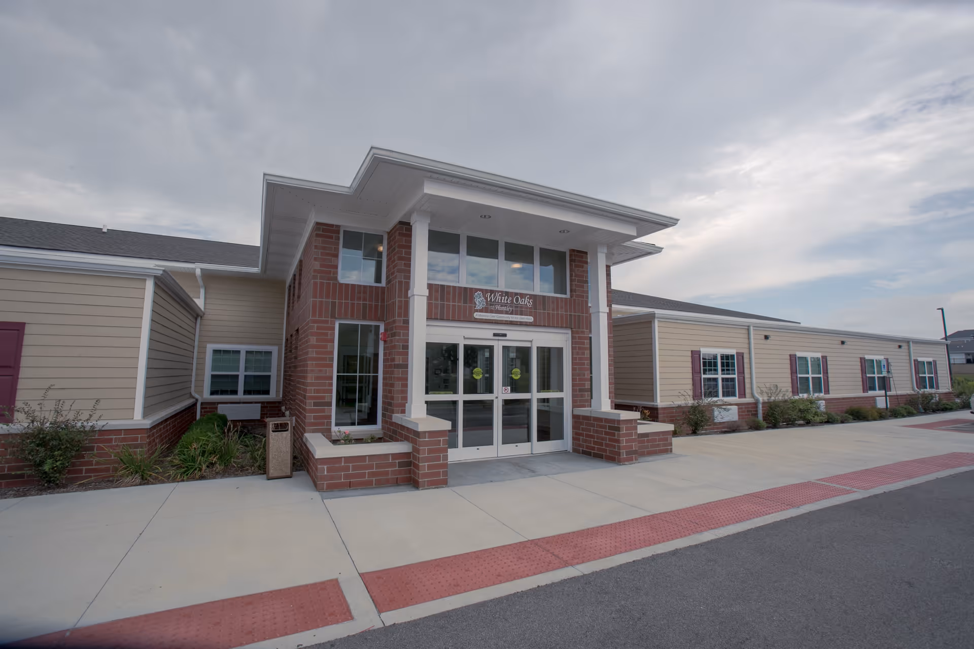 Exterior front entrance of White Oaks at Huntley facility showing a brick and beige siding building with large glass double doors under a covered entryway supported by white columns. The sky is partly cloudy.