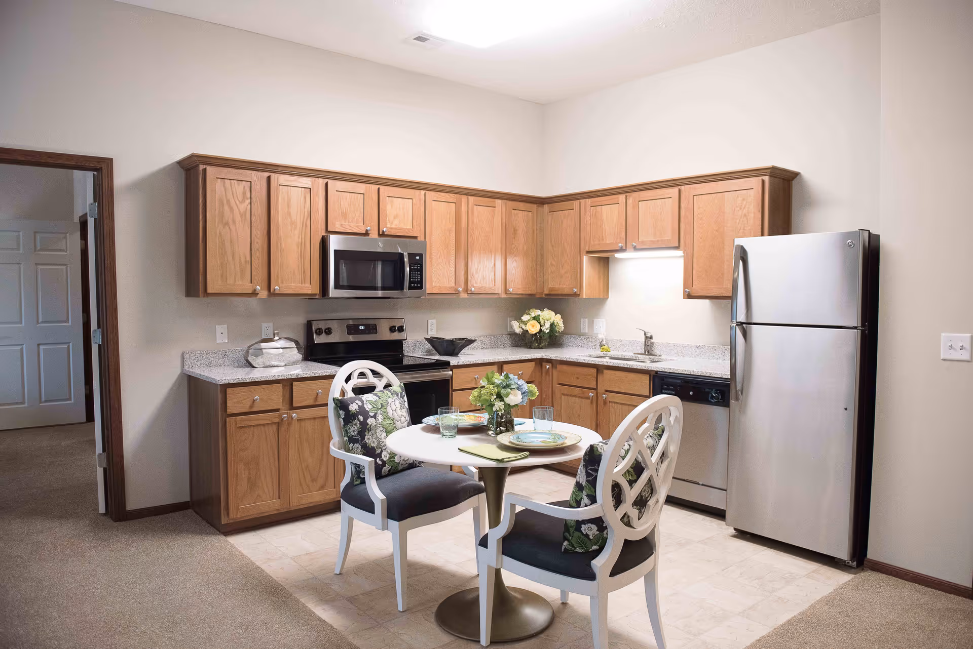 A kitchen area with wooden cabinets, a stainless steel refrigerator, microwave, stove, and dishwasher. In front of the kitchen is a small round dining table set with two chairs that have floral cushions. The table is set with plates, glasses, and a small flower arrangement. The floor transitions from carpet to tile in the kitchen area.