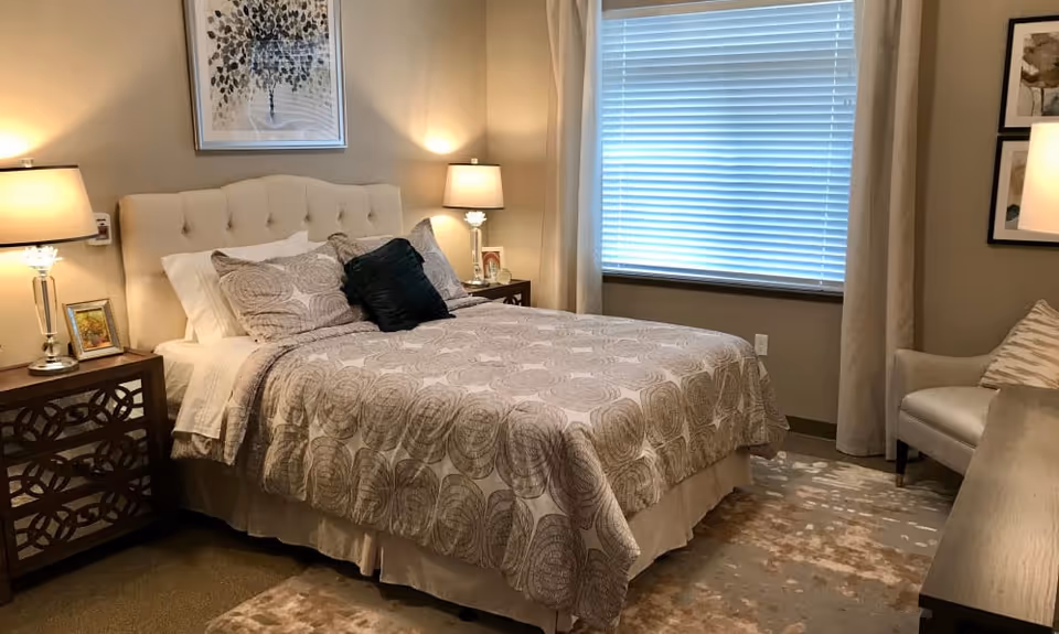 A cozy bedroom with a neatly made bed featuring patterned bedding and multiple pillows. Two bedside tables with lamps flank the bed. A window with closed blinds and curtains is on the right wall. There is a cushioned chair with a pillow and a wooden dresser in the room. The walls are decorated with framed artwork.