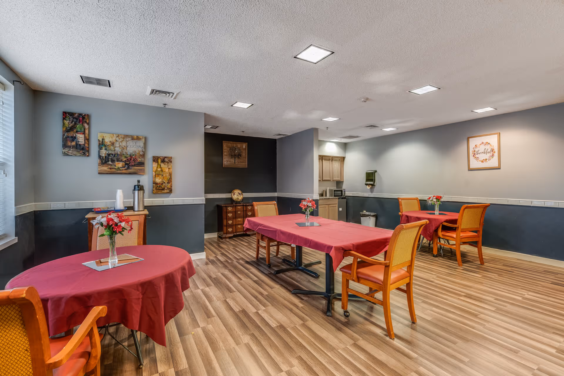Communal dining room with several tables covered in red tablecloths, wooden chairs, floral centerpieces, and artwork on the walls.