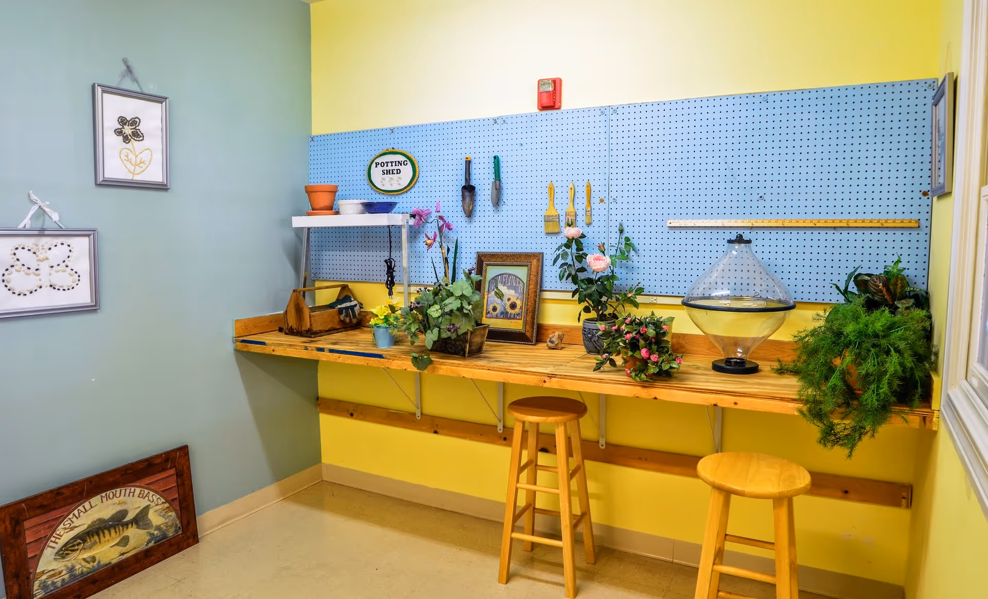 A bright potting shed area with a wooden workbench attached to yellow walls. The workbench holds various potted plants, gardening tools, and decorative items. Above the bench is a blue pegboard with hanging gardening tools and a small sign that reads 'POTTING SHED'. Two wooden stools are placed in front of the bench. The adjacent wall is painted green and has framed artwork hanging on it. The floor is light-colored and clean.