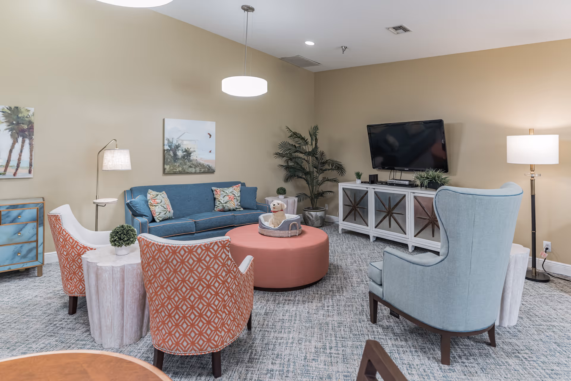 A cozy living room with a blue sofa adorned with floral pillows, two orange patterned armchairs, a light blue wingback chair, and a round pink ottoman in the center. A small dog sits in a basket on the ottoman. The room features a TV mounted on the wall above a white cabinet, a floor lamp, a potted plant, and framed artwork on beige walls.