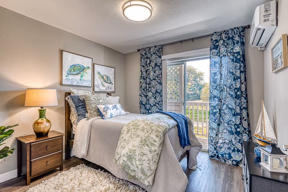 A cozy bedroom with a single bed dressed in white and green bedding, multiple pillows, and a blue throw blanket. There is a wooden nightstand with a green and brown lamp on the left side of the bed. Two framed turtle paintings hang on the wall above the bed. Blue patterned curtains frame a sliding glass door that opens to an outdoor area with greenery. A small black dresser with decorative items, including a model sailboat, is on the right side of the room. The floor is wood with a white shag rug beside the bed.