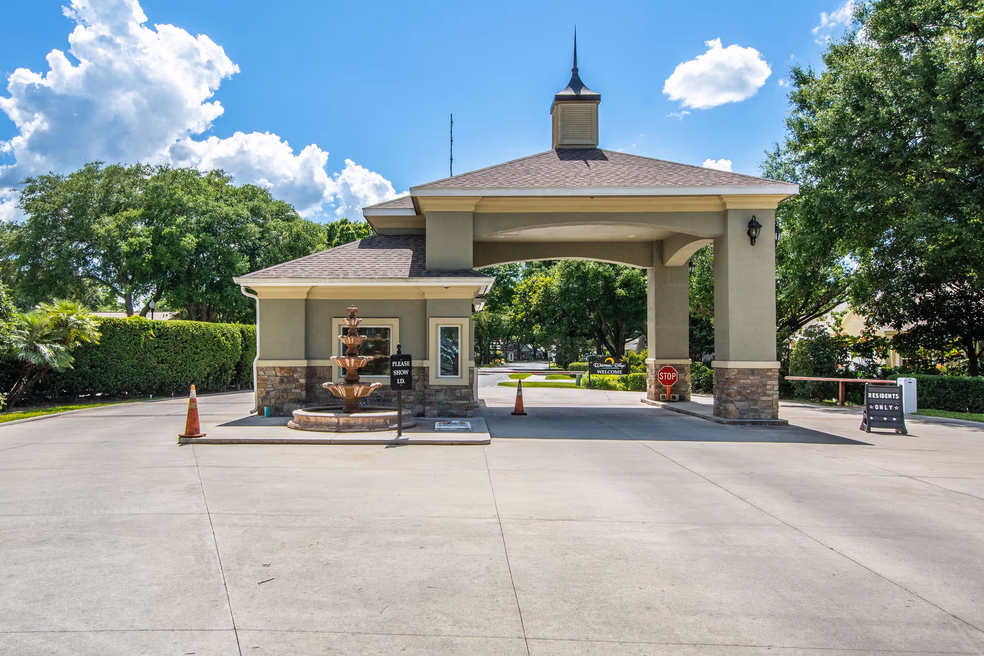 Covered entrance/guardhouse with a tiered fountain and drive-through canopy at a retirement community, surrounded by trees and signage.