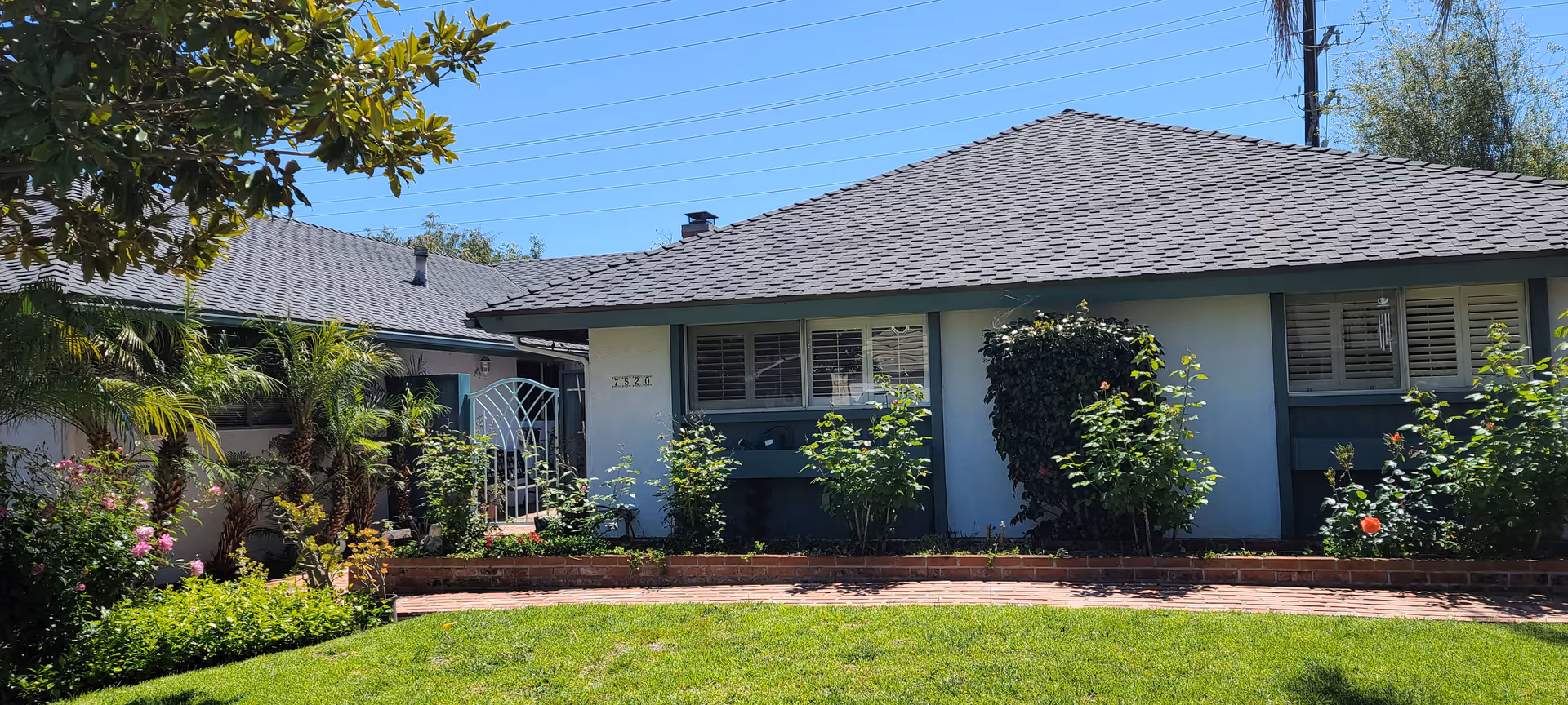 Single-story residential building with a gray shingled roof, white walls, and green trim around the windows. The front yard features a well-maintained lawn, a brick pathway, and various shrubs and flowering plants. A white metal gate is visible on the left side of the building under a clear blue sky.