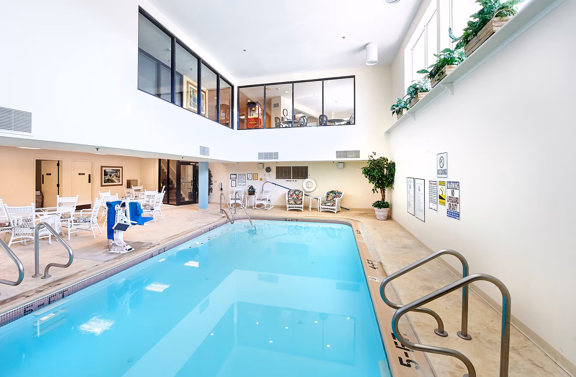 Indoor swimming pool area with clear blue water, surrounded by a beige tiled deck. There are white chairs and tables on one side, a pool lift for accessibility, and some potted plants. Large windows near the ceiling let in natural light, and there is a second-floor area with glass walls overlooking the pool.