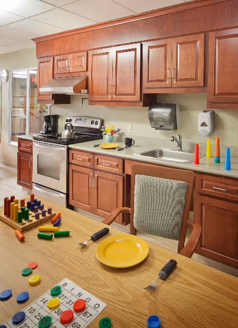 Well-lit kitchenette with wooden cabinets, a stainless steel stove and sink, and a table set with a yellow plate, utensils, colorful toys and a bingo card.