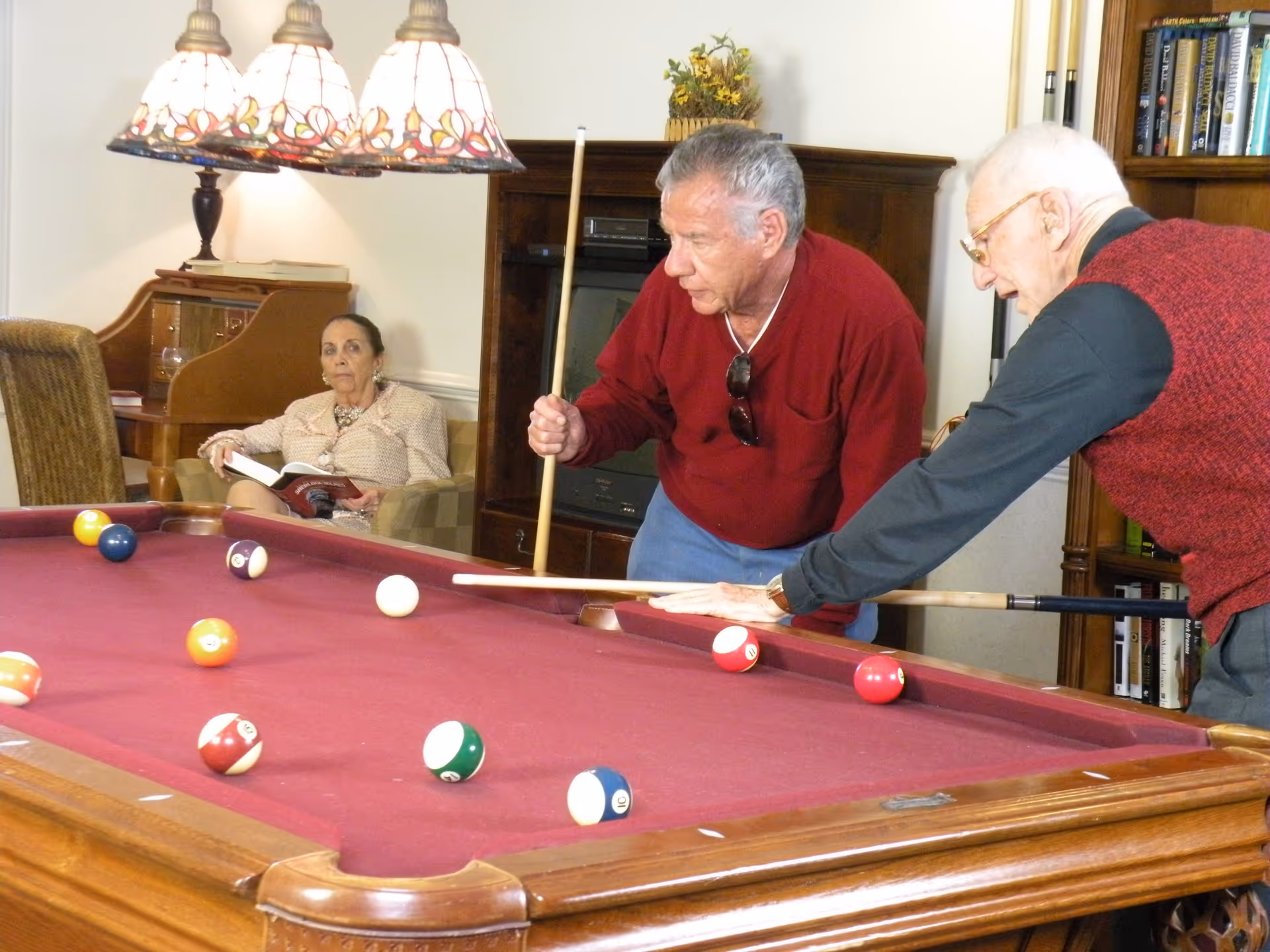 Two elderly men playing pool on a red felt pool table in a cozy room, while an elderly woman sits in the background reading a book. The room has warm lighting with a decorative hanging lamp above the pool table and wooden furniture including a bookshelf and a cabinet.