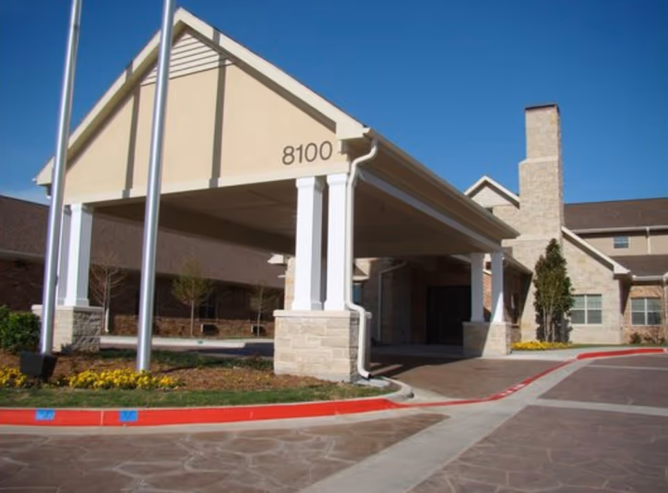 Entrance of a senior living facility with a covered drop-off area supported by white columns and stone bases. The building has beige walls, a chimney, and a clear blue sky above. The number 8100 is displayed on the front of the covered entrance. There are flower beds and small trees near the entrance.