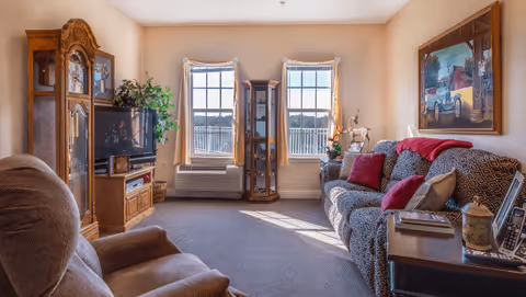 A cozy living room in a senior living facility featuring a patterned sofa with red and beige cushions, a wooden side table with books and a lamp, a recliner chair, a wooden grandfather clock, a TV on a wooden stand, two windows with light curtains letting in natural light, and a painting on the wall.