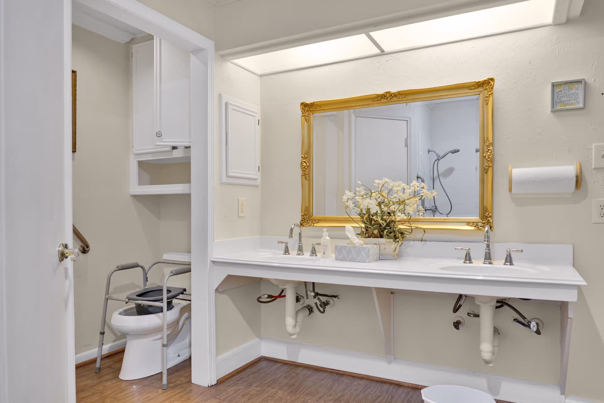 A clean and accessible bathroom featuring a double sink vanity with a large ornate gold-framed mirror above it. On the countertop, there is a decorative flower arrangement, a tissue box, and a soap dispenser. To the left, there is a toilet equipped with a safety frame for support. The bathroom has light-colored walls and wood-style flooring.
