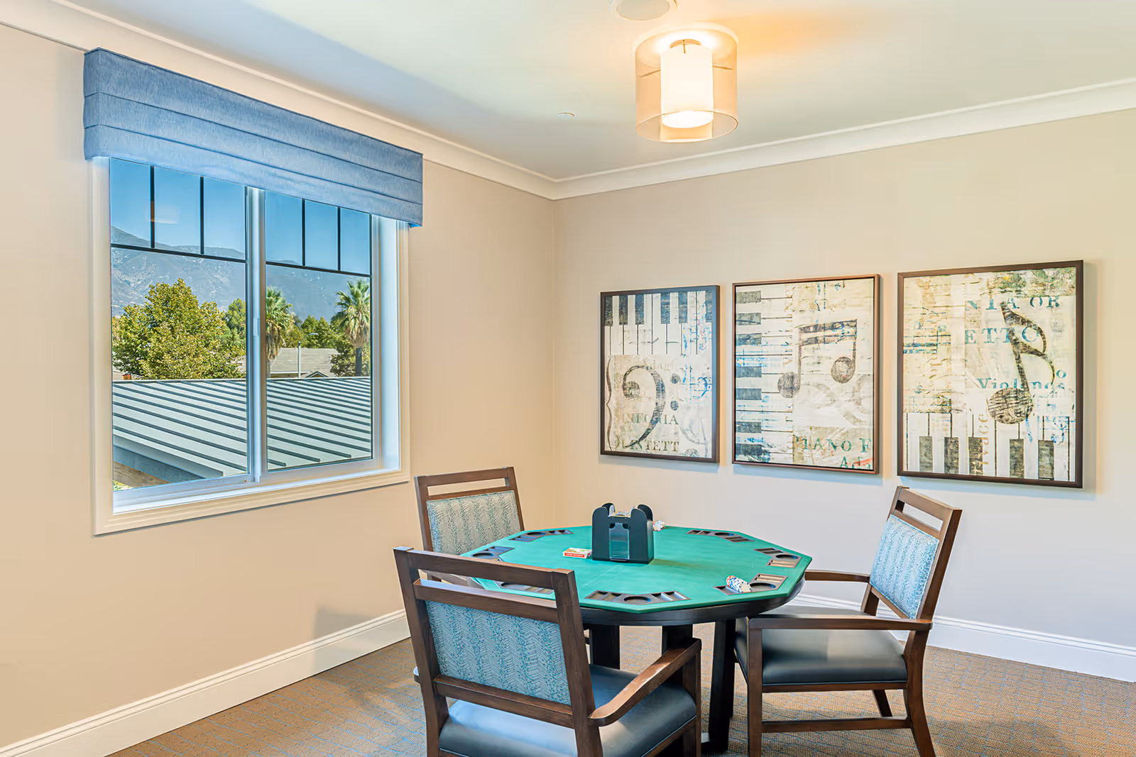 A small game room with a green octagonal card table surrounded by four wooden chairs with blue patterned cushions. On the table are poker chips and a card holder. The room has beige walls with three framed musical-themed artworks hanging on one wall. A window with a blue valance shows a view of trees, rooftops, and mountains outside. A ceiling light fixture is visible above the table.