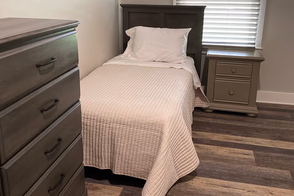 A neatly made single bed with a white quilted bedspread and a white pillow, positioned against a wall with a window covered by blinds. Next to the bed is a wooden nightstand with two drawers, and to the left is a matching wooden dresser with four drawers. The floor is covered with wood-patterned vinyl or laminate flooring.