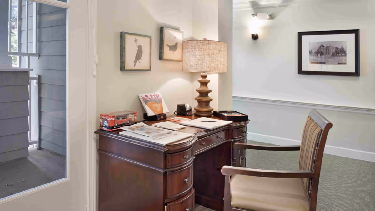 A wooden writing desk with a lamp, papers and a chair in a softly lit interior office or reception nook.