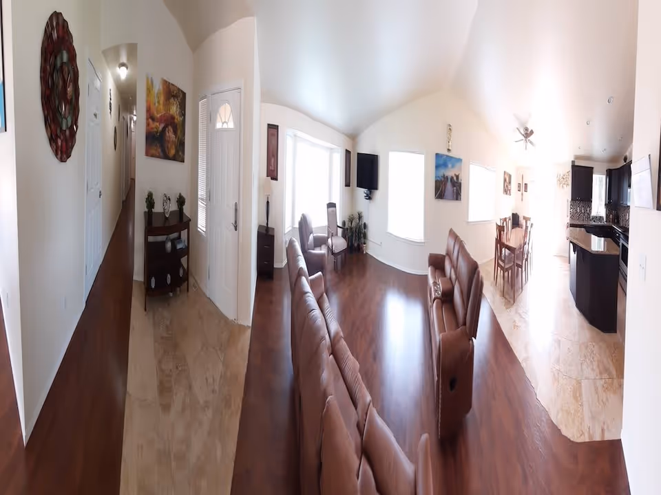 Interior view of a senior living facility showing a hallway leading to a living room with brown leather sofas, chairs, a wall-mounted TV, and large windows letting in natural light. Adjacent to the living room is a dining area with a wooden dining table and chairs, and a kitchen with dark cabinets and granite countertops.